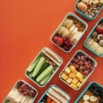 Flat lay of assorted healthy lunchboxes with fruits and nuts against vibrant orange background.